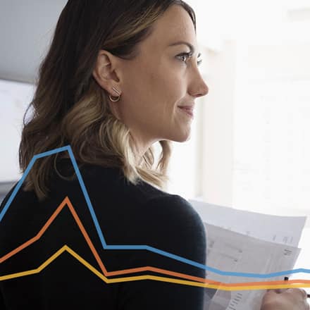 An economist reviewing paperwork at her desk with a blue, orange and yellow line graph overlay.