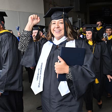 A graduate wearing a first generation stoll raising a fist and smiling while walking in a line of other graduates holding their degrees..