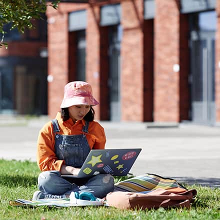 A Gen Z college student sitting in a grassy area while doing schoolwork on her laptop.