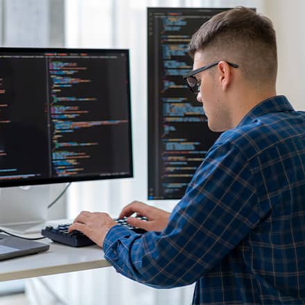 A computer science student with a laptop and two monitors of code in front of him, considering what master's degree to get.