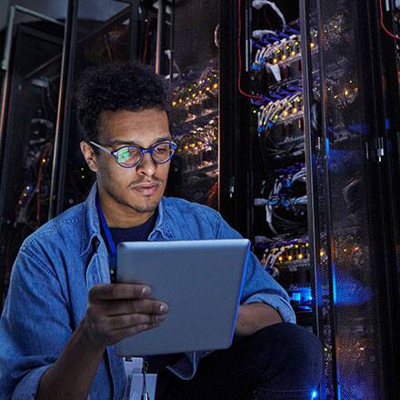 A student exploring what cyber security is, holding a tablet and standing in front of large machines at his internship.