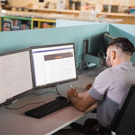 A transfer student sits in a library working on online coursework using two computer monitors.