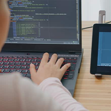 A person coding at a laptop while referring to a tablet featuring more code and a video call in the corner of the screen.