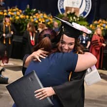 An SNHU graduate dressed in cap and gown hugging an advisor.