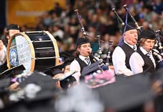 A group of people playing bagpipes and drums processing into the SNHU Arena during a Commencement ceremony.