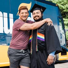 Jesús Suárez wearing his cap and gown with his partner John in front of the Southern New Hampshire University bus