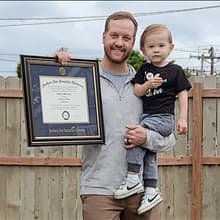 Wyatt Martensen outside holding his SNHU diploma in one hand and his young son in the other. 