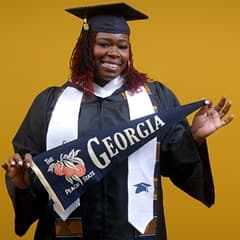 Candace Boyer, a 2025 SNHU graduate who earned her bachelor's in psychology wearing her cap and gown in front of a yellow background and holding a Georgia state flag.