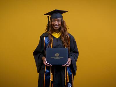 Chantel Hutchison, a 2025 SNHU graduate who earned her bachelor's in public health, holding her diploma and wearing her cap and gown in front of a yellow background.