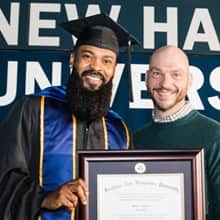An SNHU graduate wearing a cap and gown, holding a framed diploma and standing beside a supporter.