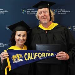 2024 SNHU graduates, David and Irina Roach, who earned their master's in cybersecurity, holding a California state flag, wearing their caps and gowns in front of a blue background.