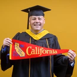David Wallace, a 2025 SNHU graduate who earned his bachelors in business administration wearing his cap and gown in front of a yellow background holding a California state flag.