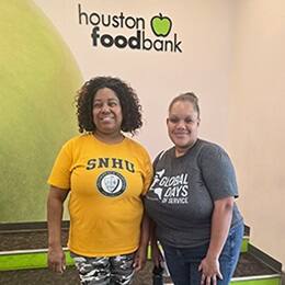Two volunteers standing and smiling beneath a Houston Food Bank sign, wearing shirts that read “SNHU” and “Global Days of Service.”