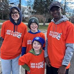 Four young volunteers wearing matching orange “Give a Little Feed a Lot” T-shirts smiling outdoors during a community service event.