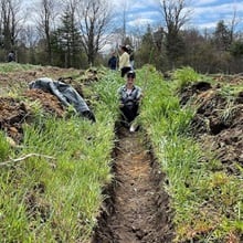 An SNHU staff member holding gardening equipment and sitting on a hill in the New Hampshire Food Bank production garden.