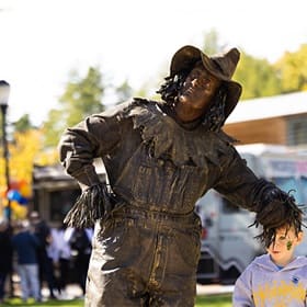 A Homecoming Street Fair performer dressed like a scarecrow with metallic paint, pretending to be a statue.
