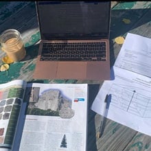 A laptop, textbook, pen, paper and drink resting on a makeshift outdoor desk.