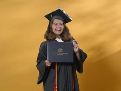 Adrian Henares, who earned her online bachelor's in communication from SNHU in 2024, wearing her cap and gown and holding her diploma in front of a yellow backdrop.