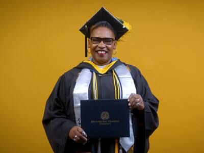 Angela Charlton, who earned her bachelor's in computer information systems from SNHU in 2025, wearing her cap and gown and holding her diploma in front of a yellow backdrop.