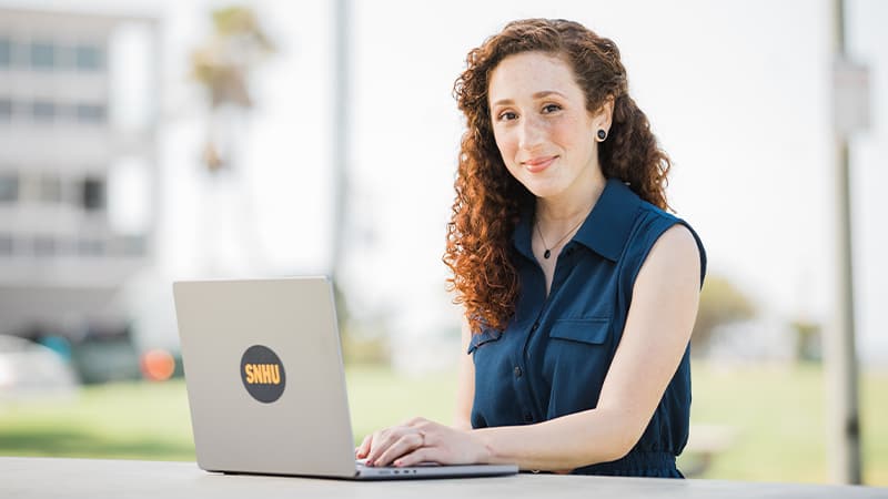 Mariel Embry, who earned her online degree from SNHU in 2022, wearing a sleeveless blue shirt and sitting at an outdoor table with an open laptop.