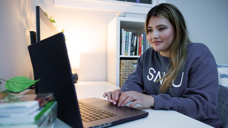 Juliana Zambrano, who earned her online degree from SNHU in 2024, wearing an SNHU sweatshirt and sitting at a desk typing on her laptop.