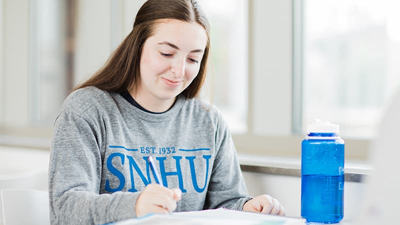 A female online college student wearing a grey SNHU sweatshirt writing in a notebook with a blue water bottle next to her.