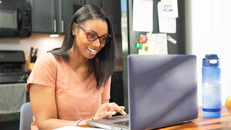 Minda Thurman, who earned her online bachelor's in public health from SNHU in 2023, wearing glasses and typing on her laptop at her kitchen table.