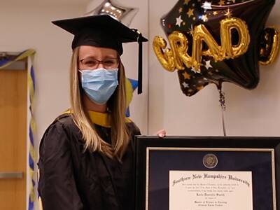 Katie Smith, who earned her MSN from SNHU in 2021, wearing her cap and gown and holding her framed diploma with a "Grad" balloon in the background.