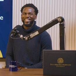 Kitrick Bell, a 2025 SNHU graduate who earned his master's in information technology with a concentration in software application development, smiling behind f a microphone and his diploma.
