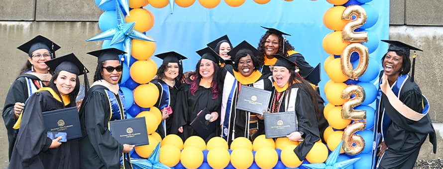 A group of SNHU graduates holding their diplomas and standing in and around a blue and yellow balloon frame.