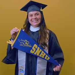 Paola Andrea Gaona Salamanca, a 2026 SNHU graduate who earned her bachelor's in business administration wearing her cap and gown and holding a SNHU flag in front of a yellow background.