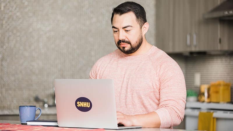Jesus Suarez, who earned his BA in Graphic Design and Media Arts in 2021, working on a laptop in his kitchen.