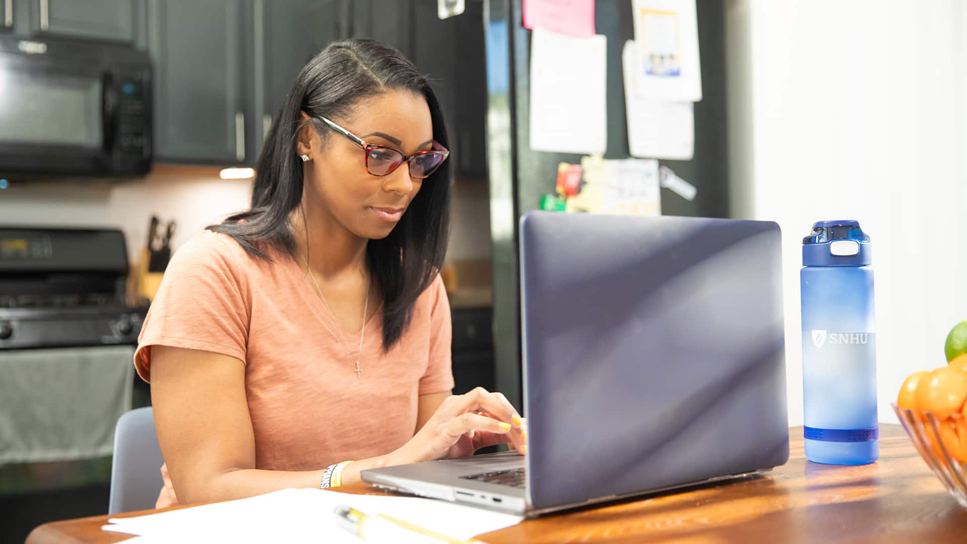 Minda Thurman, who earned her BS in public health, wearing glasses and working on her laptop in her kitchen with an SNHU water bottle on the table beside her.