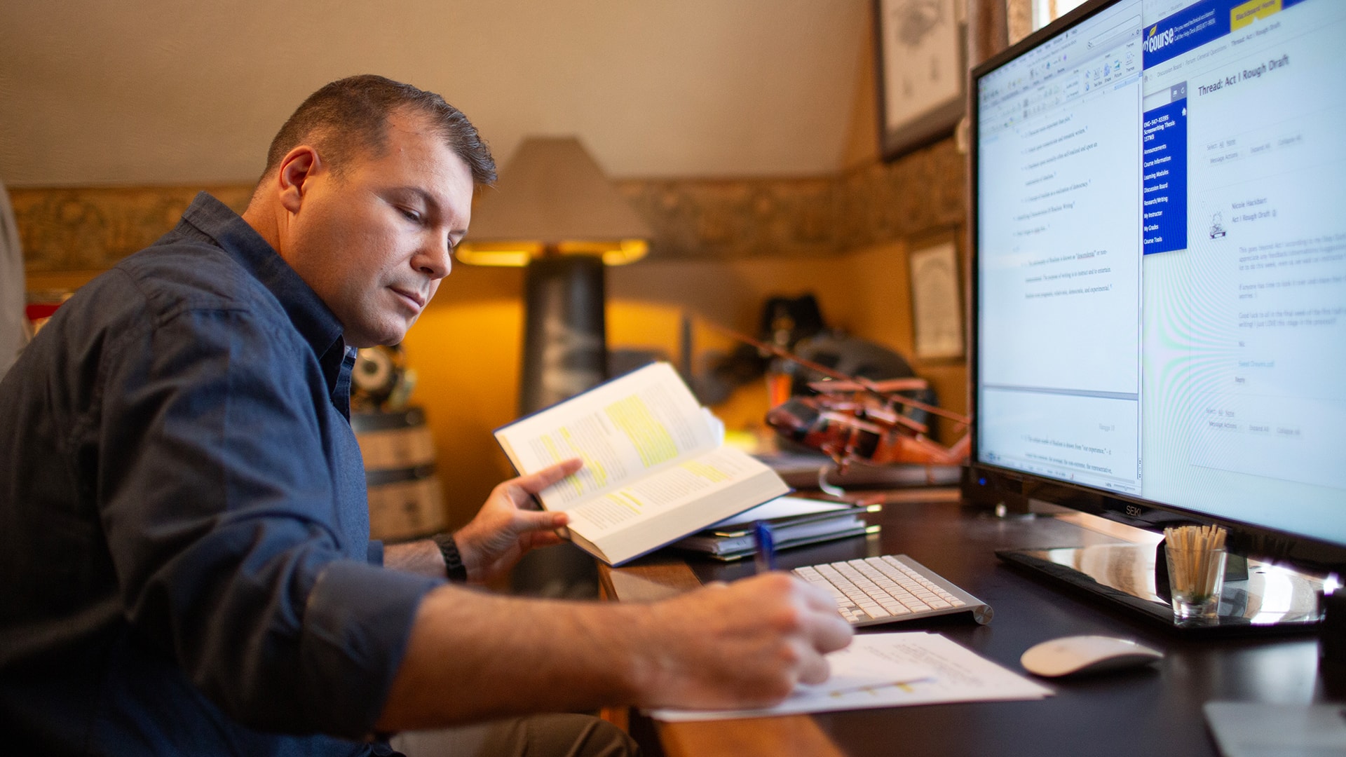Michael Hangge, an SNHU alum, at the desk in his home office in front of a computer monitor, holding open a text book with some passages highlighted and taking notes on a piece of paper.