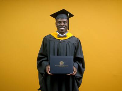Jaiwaun Haggerty, who earned his online master's in sport management from SNHU in 2025, wearing his cap, gown and a bowtie holding his diploma in front of a yellow backdrop.