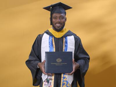 Jerron Trice, who earned his bachelor's in business administration from SNHU in 2024, wearing his cap and gown and holding his diploma in front of a yellow backdrop.