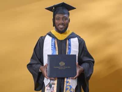 Jerron Trice, who earned his bachelor's in business administration from SNHU in 2024, wearing his cap and gown and holding his diploma in front of a yellow backdrop.