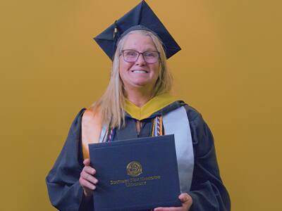 Kelly Pilotte ’25, who earned her online bachelor's in Business Administration with a concentration in Nonprofit Management, wearing her cap and gown and holding her diploma in front of a yellow backdrop.