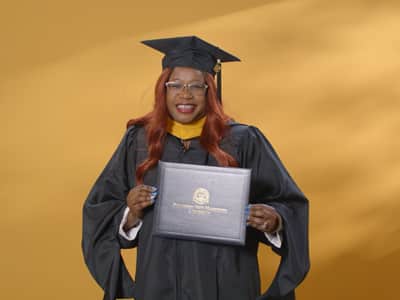 Lilian Ongaki, who earned her master's in healthcare administration in 2024, wearing her cap and gown and holding her diploma in front of a yellow backdrop.