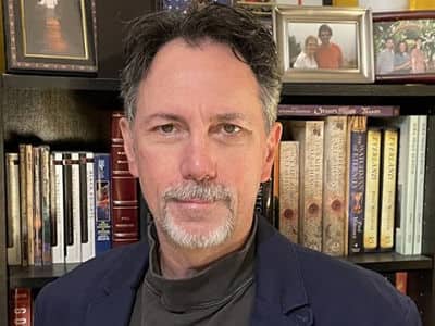Paul Witcover, associate dean of SNHU's online liberal arts program, standing in front of bookcase with framed pictures on top.