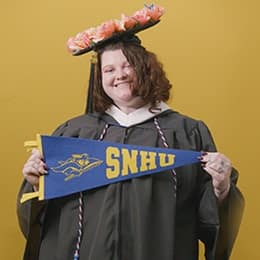Shelbie Lynne Ayles, a 2025 graduate who earned her bachelor’s in psychology with a minor in sociology from SNHU wearing her cap and gown holding a SNHU flag in front of a yellow background.