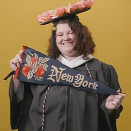 Shelbie Lynne Ayles, a 2025 graduate who earned her bachelor’s in psychology with a minor in sociology from SNHU wearing her cap and gown holding a New York flag in front of a yellow background.