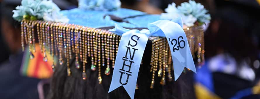 A decorated graduation cap featuring blue flowers, shimmering beads, and two ribbons that read SNHU and ‘26.