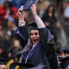 A graduate wearing a cap and gown, holding her diploma cover up over her head