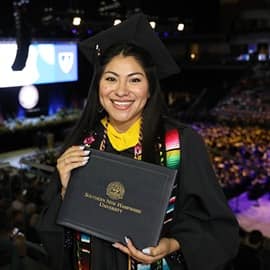 Eliana Cornejo holding an SNHU diploma cover and wearing a graduation cap, gown, cords and stole.