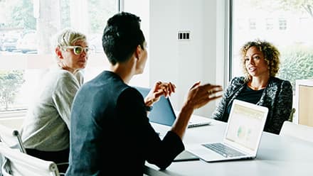 Three coworkers sitting around a table at a meeting with their laptops open for their job they got with their business degree.