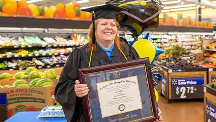 Carol D'Anna in her cap and gown holding her diploma, standing in the center of Walmart produce section 