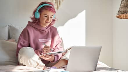 A person wearing a pink sweatshirt and teal headphones, researching on a laptop which graduate program to choose.
