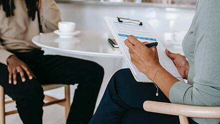 A clinical mental health counselor taking notes on a clipboard while talking to a client.