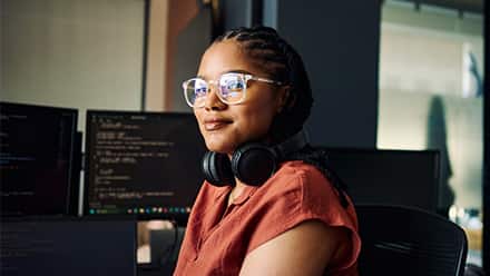 A master's in computer science student with over-ear headphones around her neck, sitting at a desk with code on the monitor.